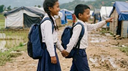 Lachende nepalesische Schulkinder in Seitenansicht mit blauen Schulrucksäcken auf dem Weg durch das Krisengebiet Nuwakot