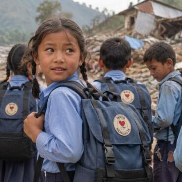 KI-generierte Illustration: Nepalische Schulkinder tragen blau-schwarze Rucksäcke der Maya Nepalhilfe vor zerstörten Häusern nach der Flutkatastrophe in Nuwakot.